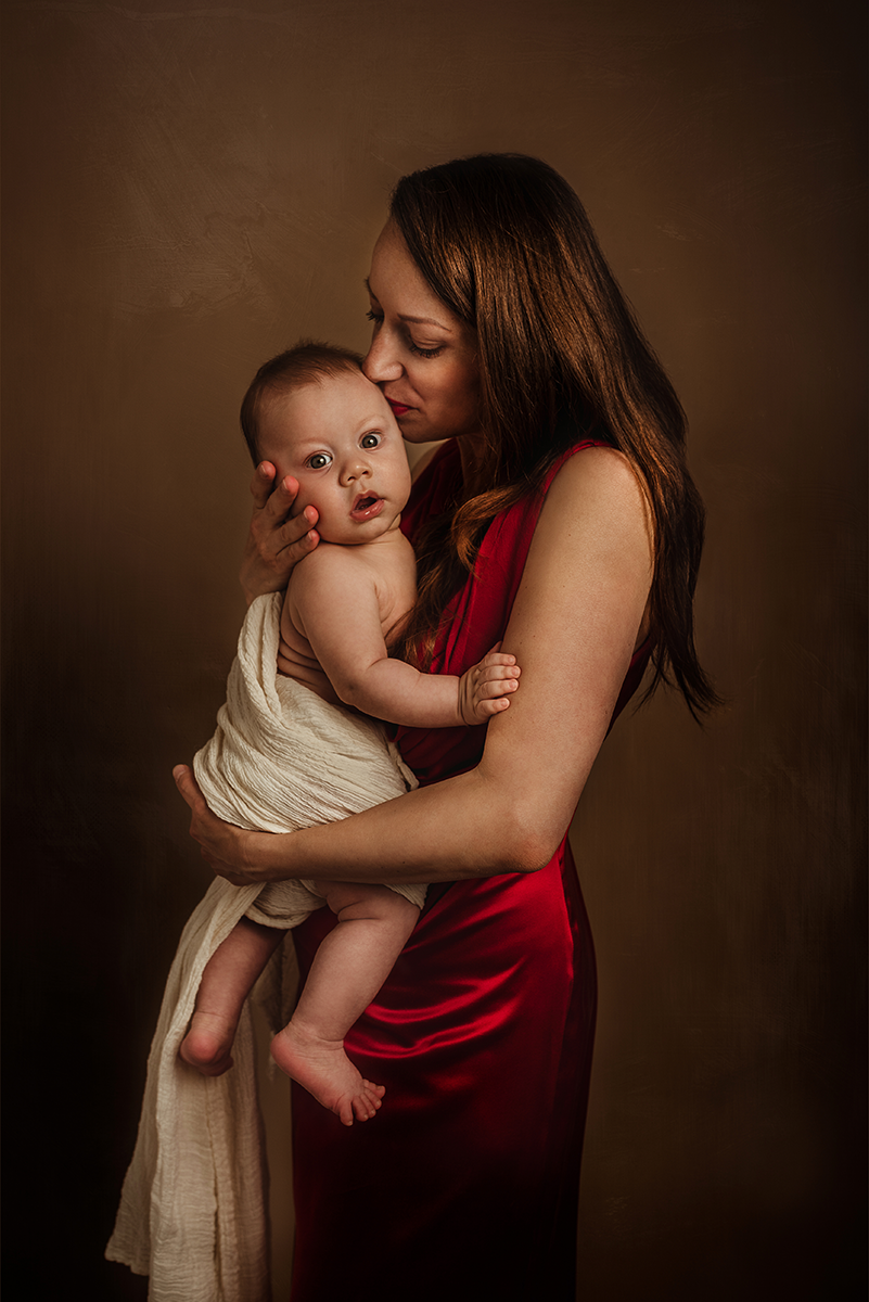 Ritratto madre in vestito elegante che bacia il suo bambino tenendolo in braccio su sfondo marrone